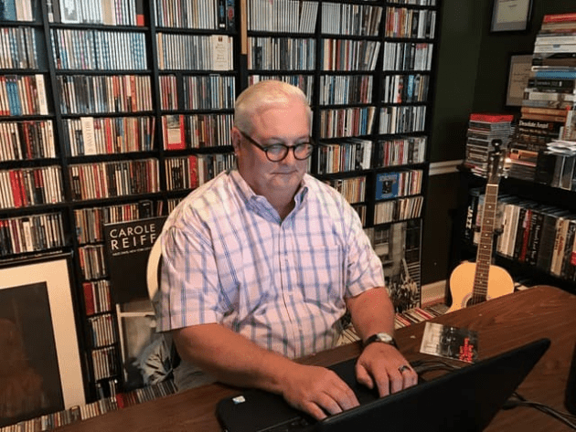 Michael Bailey writing on a laptop with music collection on shelves behind him.